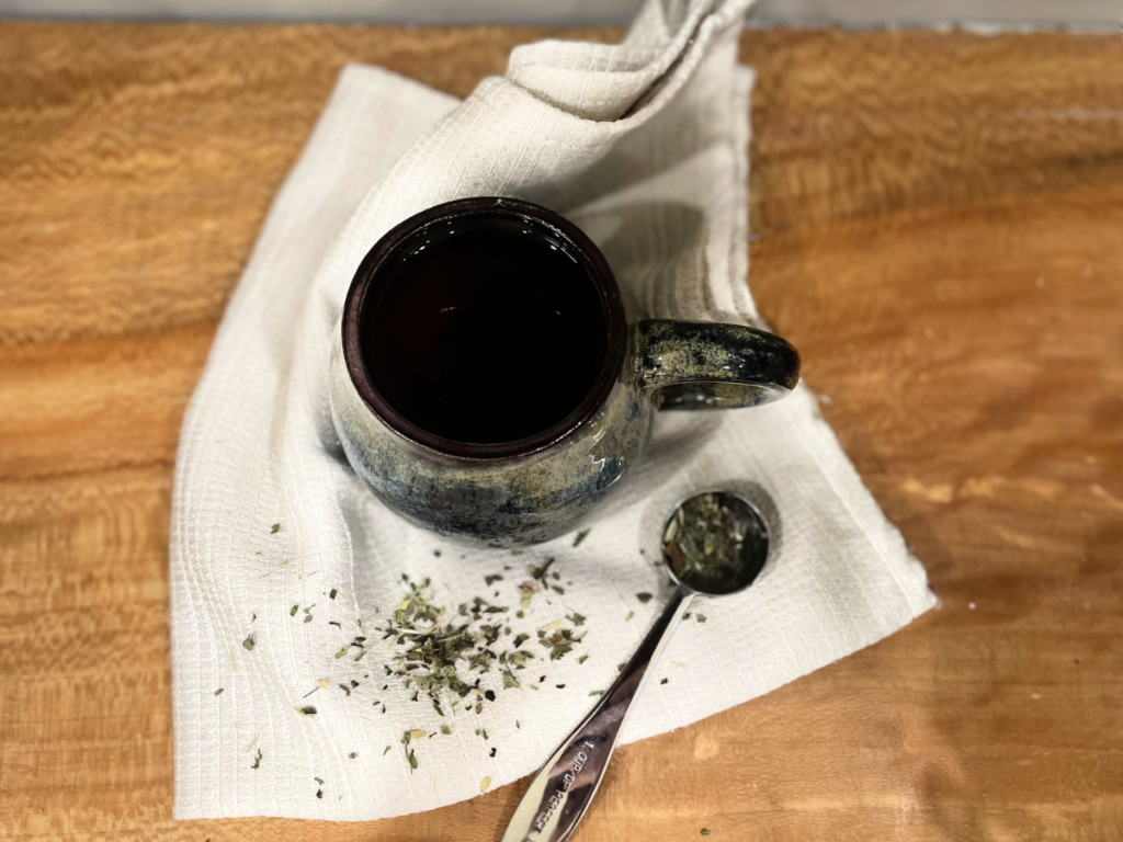 Picture of tea in a mug on a white napkin next to a teaspoon full of dry tea leaves.