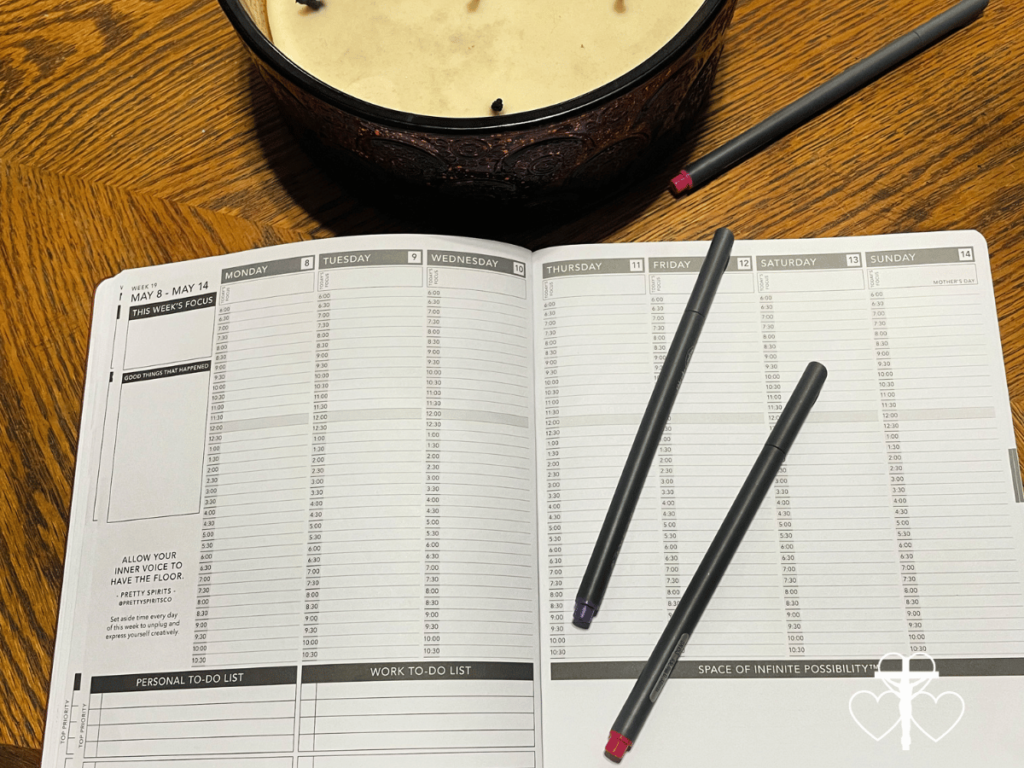 Picture of a planner and candle with several pens to write with on a wooden table.