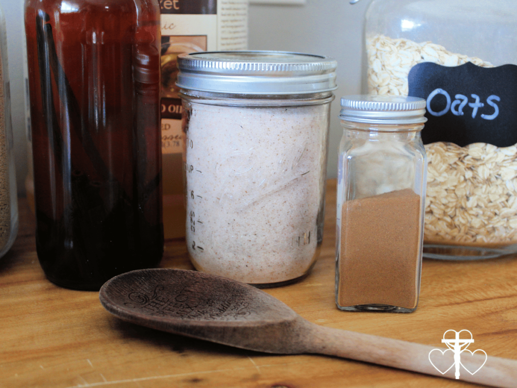 Picture of a jar of salt, oats, cinnamon, and vanilla with a wooden spoon on a wooden counter.