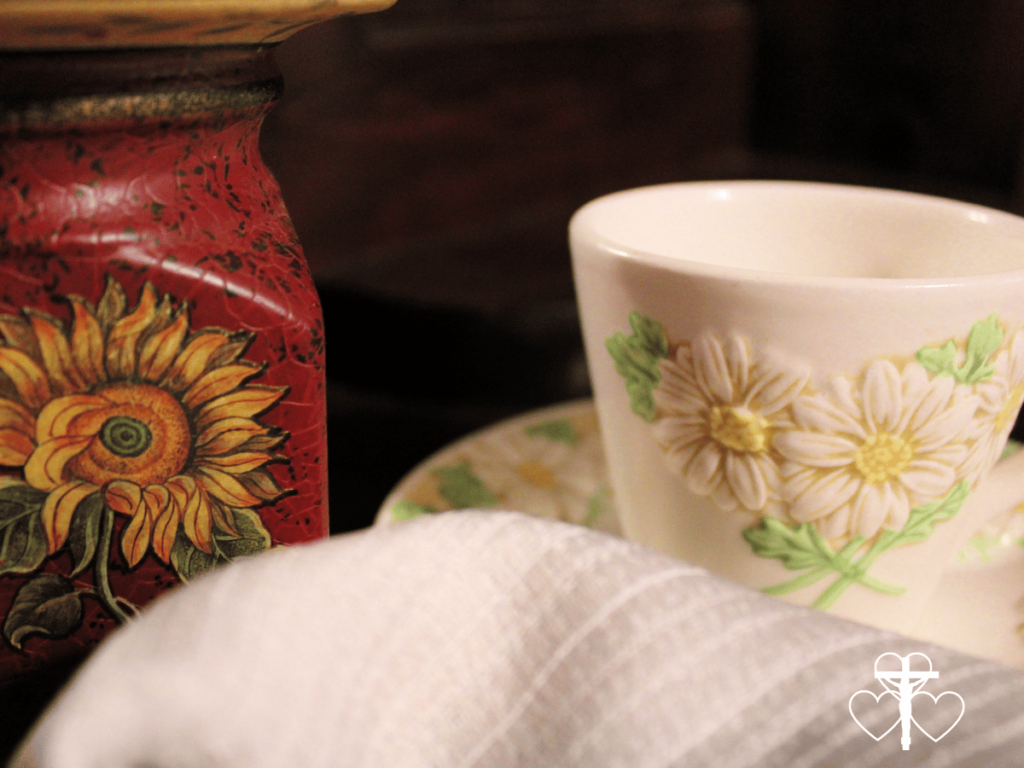 Picture of a white tea cup and saucer etched with daisies sitting next to a sunflower painted candle stand.