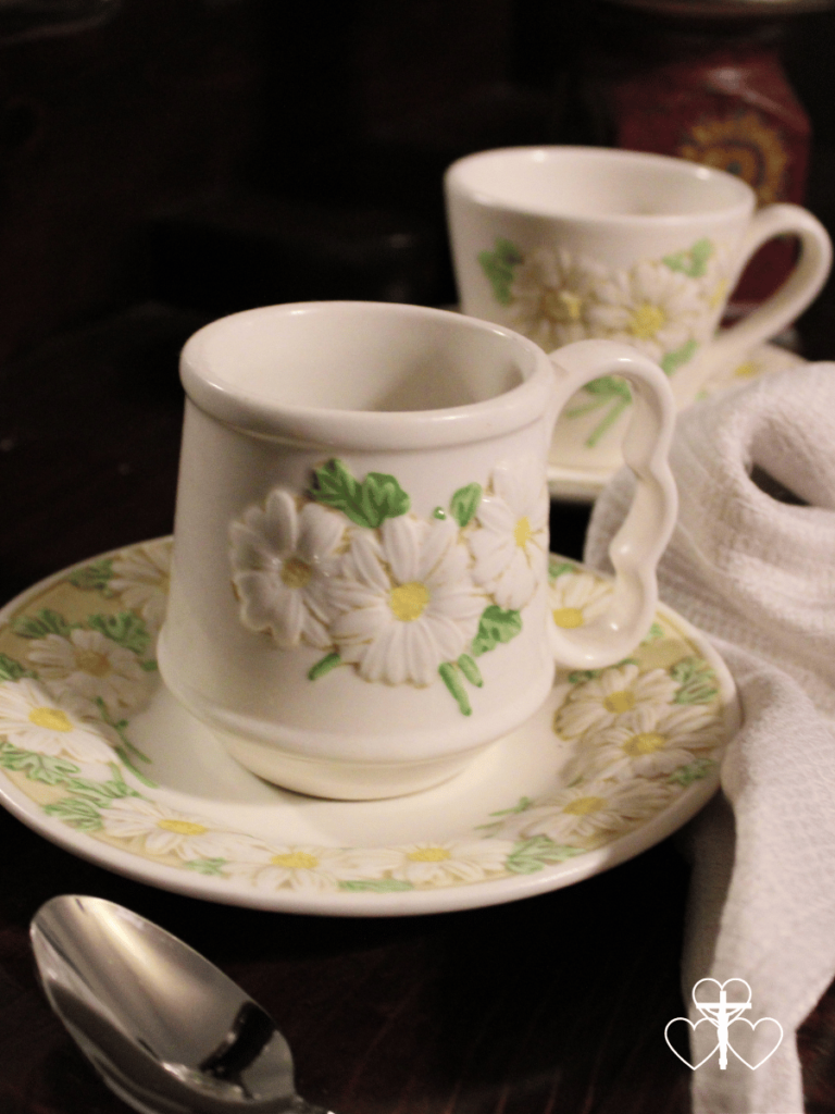 Picture of sculpted daisy coffee mug and tea cup sitting on a wooden table with a cream napkin surrounding them.