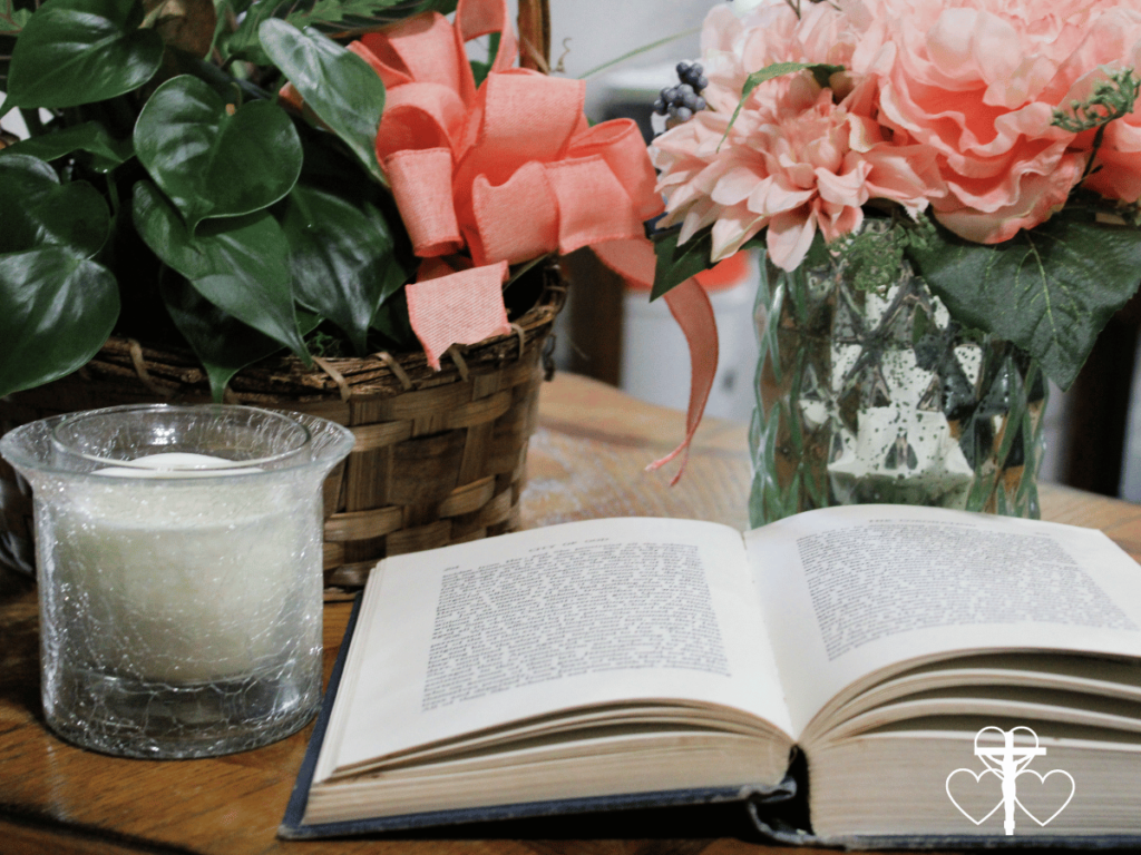 Picture of an open book on a table sitting next to a candle in a glass votive with flower arrangements behind them.